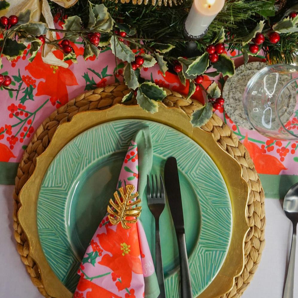 Mayajoy Christmas poinsettia cotton napkin styled on Christmas table scape with sage green plate, gold charger plates and Christmas poinsettia table runner with holly, red berries and candles.