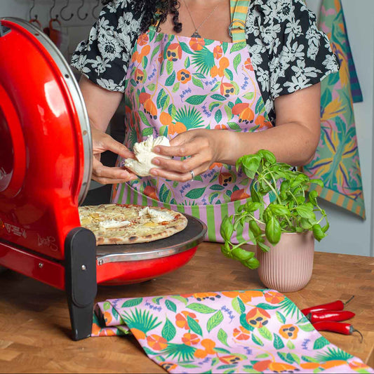Woman in Ackee stripe cotton apron, with hand illustrated Ackee print with pink background, green leaves and orange Ackee and lilac thyme leaves, making a pizza with green pizza oven 