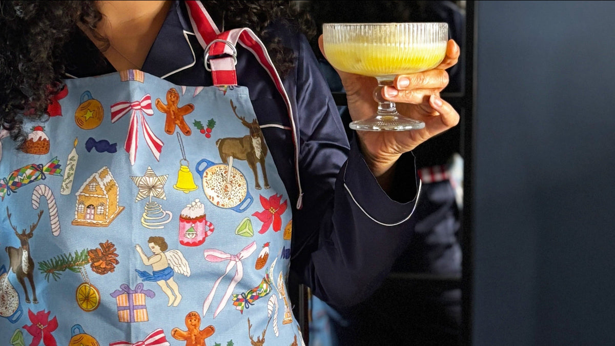 Woman wearing festive apron with blue background and hand drawn Christmas motifs, with red and pink candy stripe large pocket and neck tie, holding festive snowball cocktail 