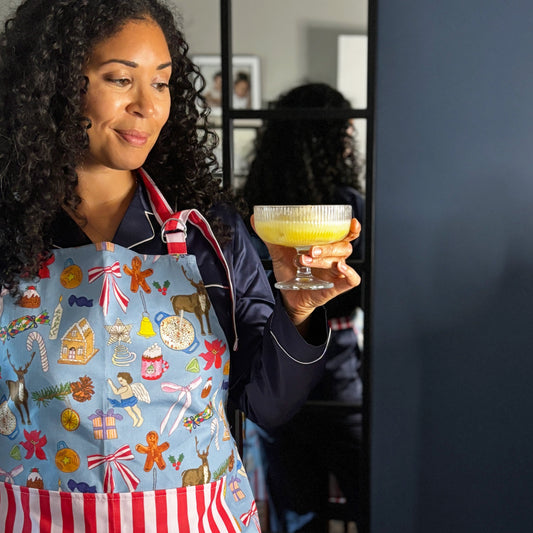 Woman wearing festive apron with blue background and hand drawn Christmas motifs, with red and pink candy stripe large pocket and neck tie, holding festive snowball cocktail 