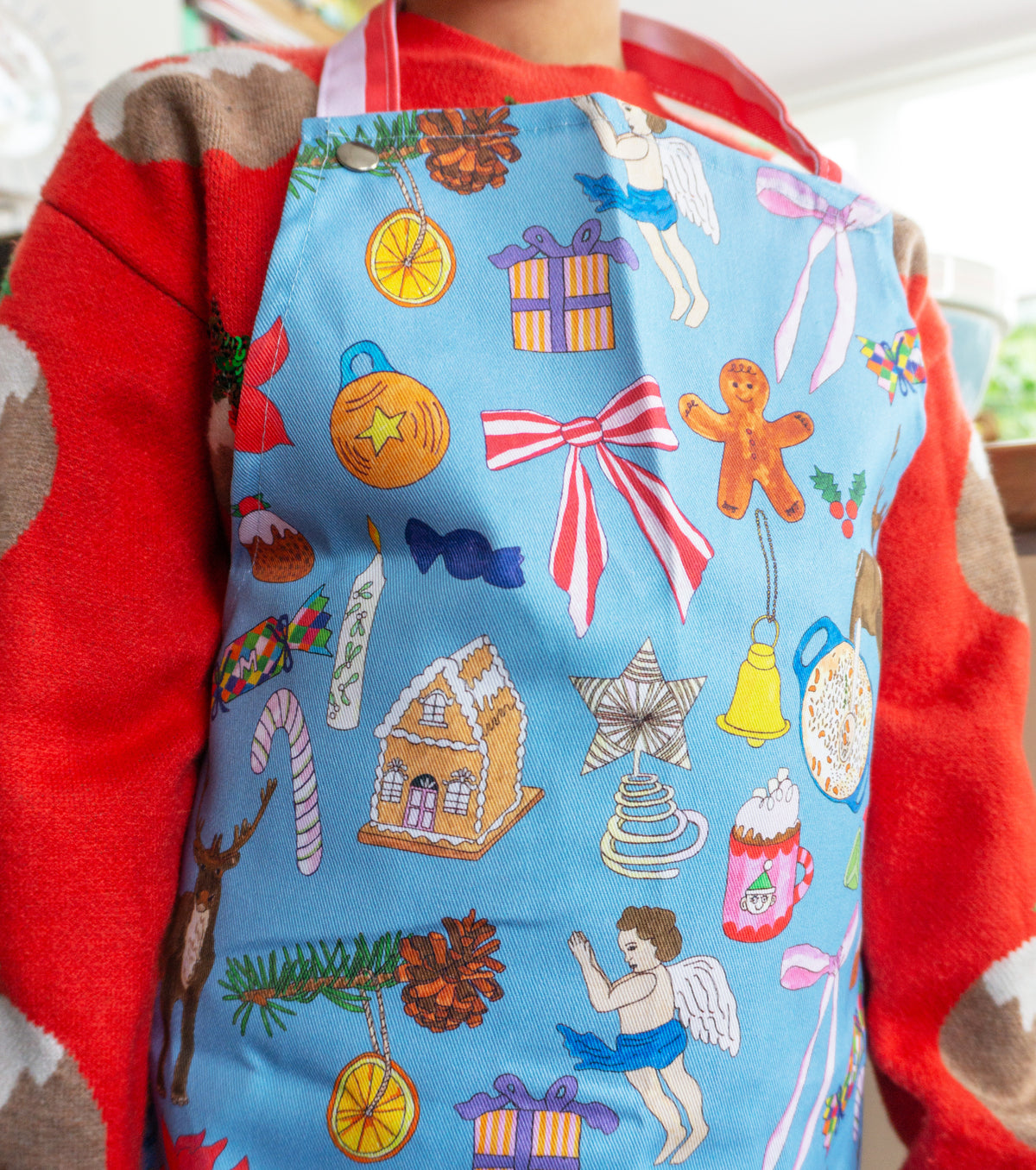 Lifestyle image of child wearing festive apron in blue with hand illustrated Christmas motifs with red and pink candy stripes to neck 