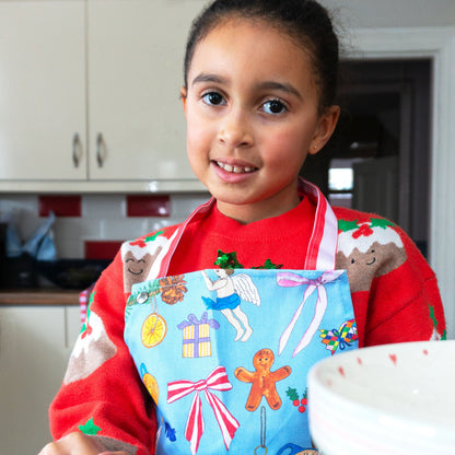 Child wearing hand illustrated Christmas apron in blue, with festive motifs and pink and red candy stripes to neck