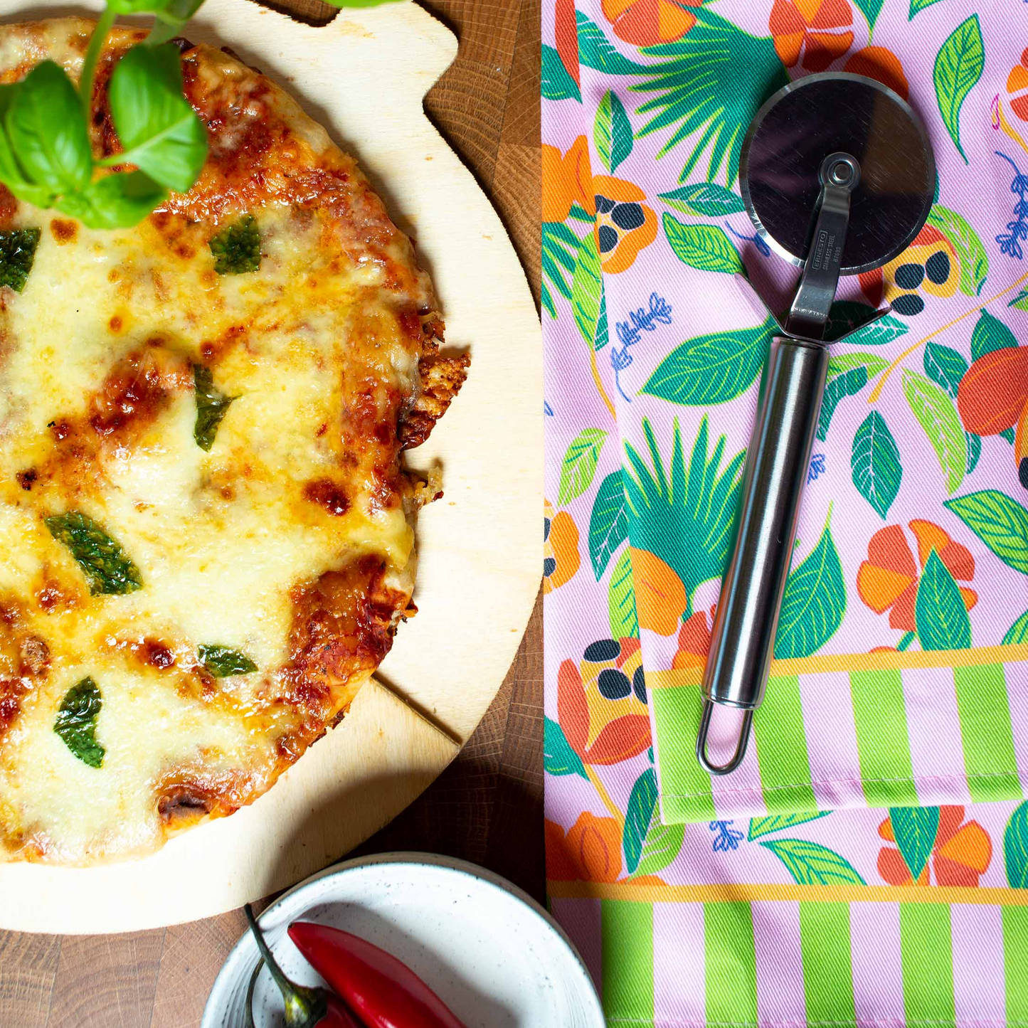 Colourful cotton tea towel in hand illustrated ackee print with pink background, green leaves, orange ackee and lilac thyme leaves, styled next to pizza and red chillies 