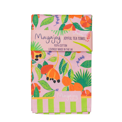 Mayajoy colourful cotton tea towel with hand illustrated ackee design with pink background, green leaves and orange ackee with lilac thyme leaves folded with coral belly band and words “joyful tea towel”