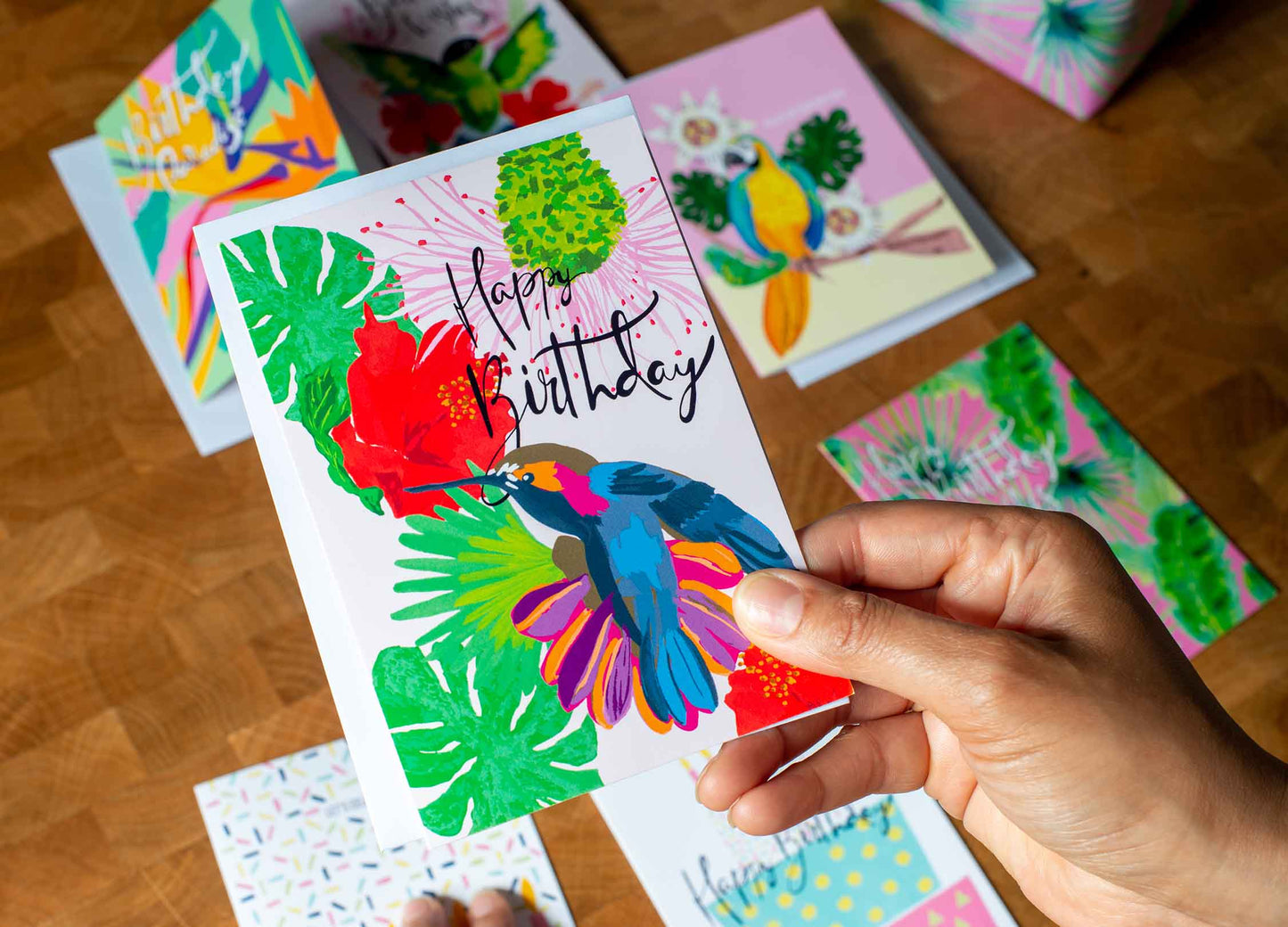 Hand illustrated Jamaican hummingbird card in pink, green, blue, orange and red with hibiscus flowers and hand calligraphy “happy birthday”, styled with hands holding card and other greeting cards I’m background,  by Mayajoy .