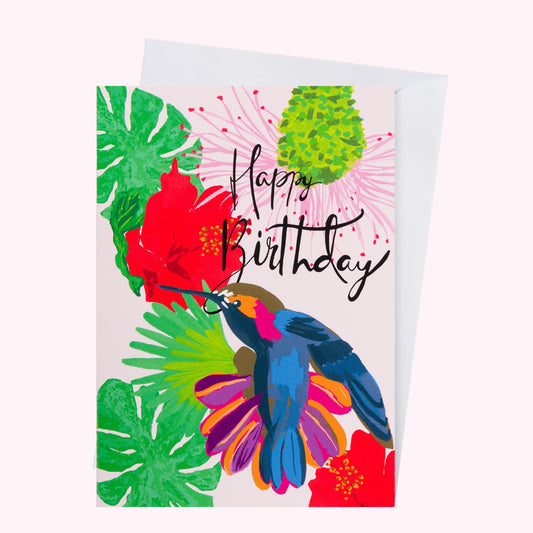 Hand illustrated Jamaican hummingbird card with hibiscus flowers and tropical foliage in pink, green, red, orange and blue with hand calligraphy words “happy Birthday”