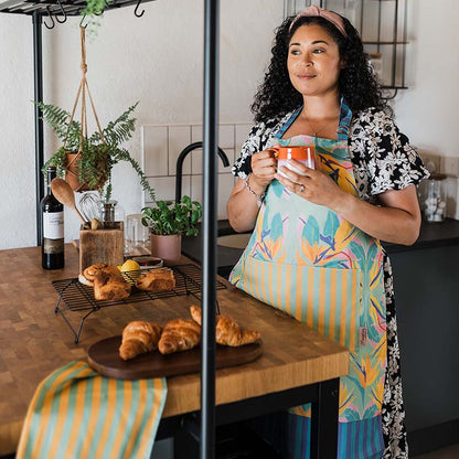 Woman in kitchen wearing cotton apron in birds of paradise design, featuring green, pink, orange and purple design with striped pocket and ties