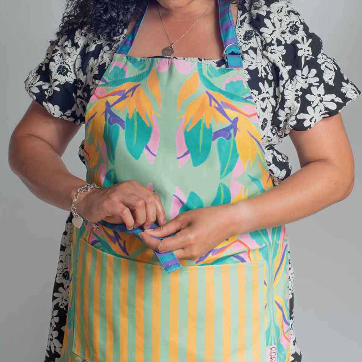 Woman wearing an apron with colourful birds of paradise in green, orange, pink and purple with stripy neck strap and ties 