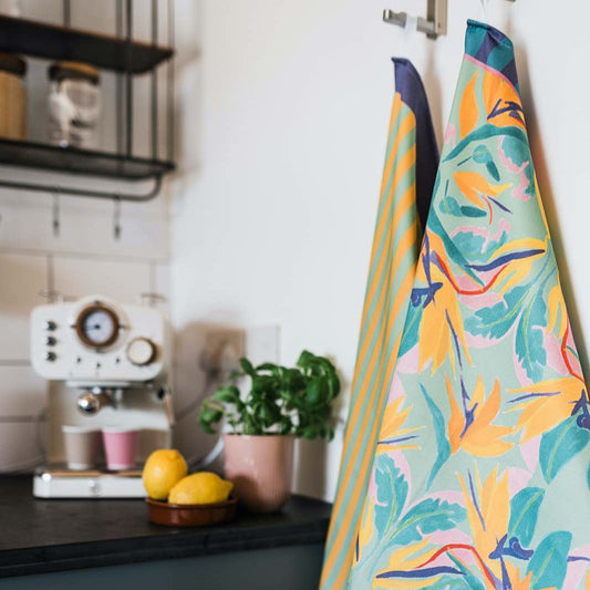 Mayajoy duo colourful cotton tea towels in paradise print, featuring orange birds of paradise flower, green, pink and purple and paradise stripe in green and yellow stripe with purple edge, both hanging on hook in kitchen