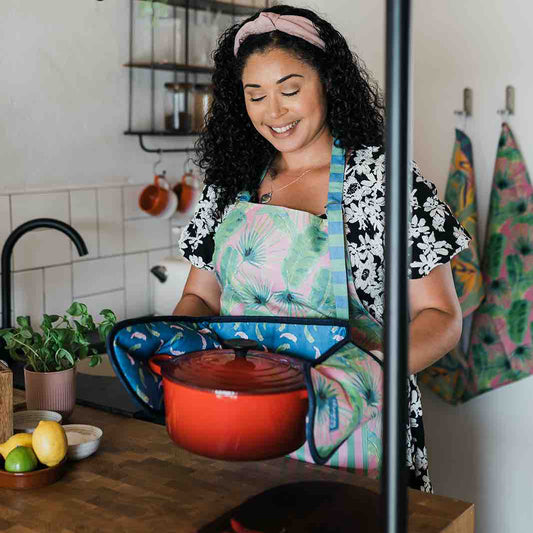 Woman in kitchen wearing pink palm stripe apron in tropical foliage pattern in pink and green, with stripy pocket and ties, holding le creuset and oven gloves 