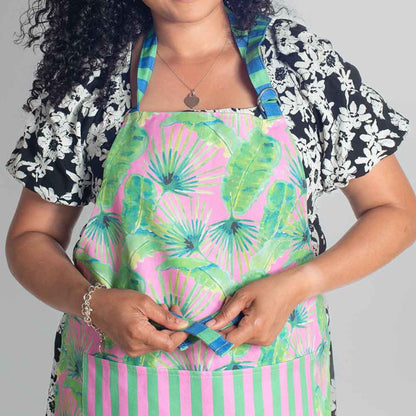 Woman wearing pink palm colourful cotton apron tying at waist, with pink and green tropical foliage design with stripe pocket and ties 