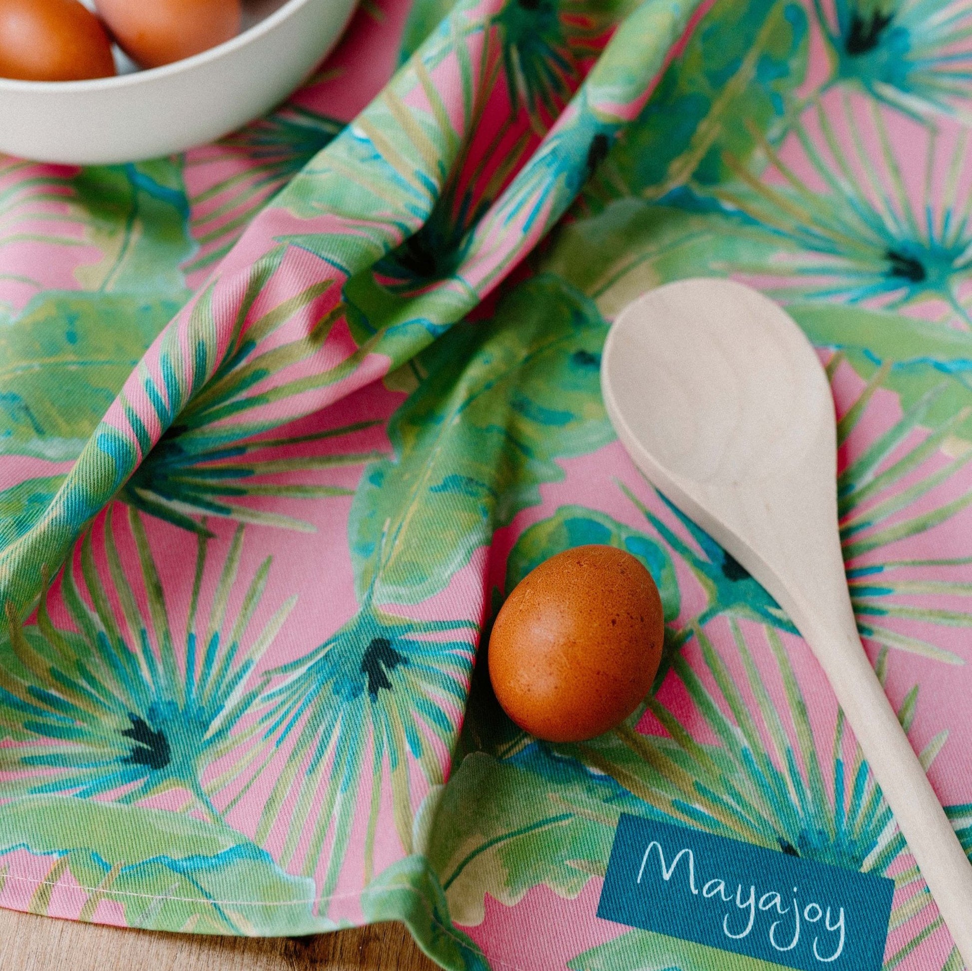 Colourful tea towel in pink palm with green, blue and pink hand illustrated foliage by Mayajoy, styled with eggs and wooden mixing spoon 